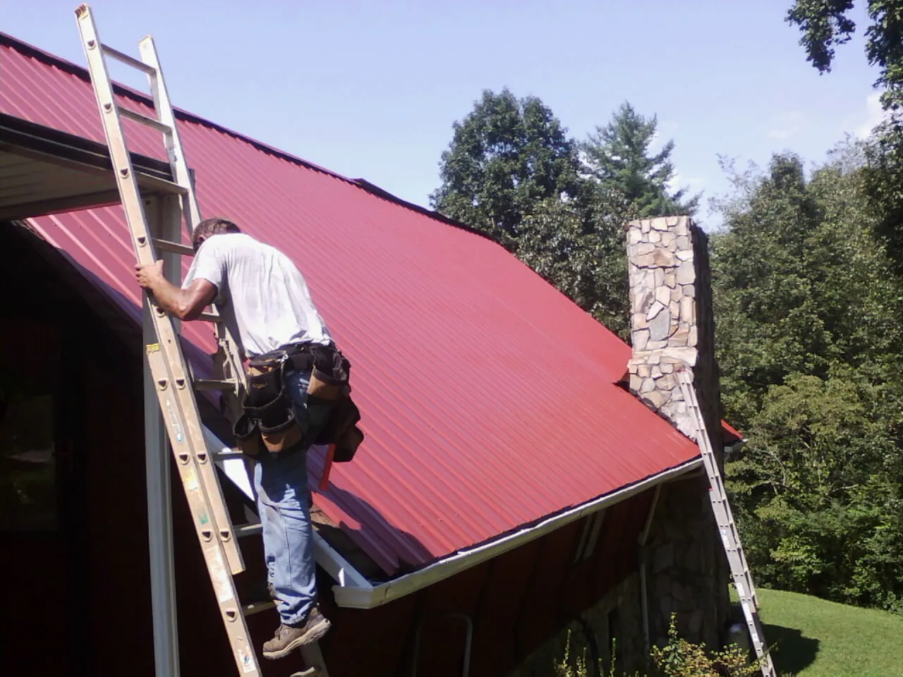 Roofer on a ladder beside a red metal roof and stone chimney — Soffit & Fascia Repair by Roofing Brothers in Albert